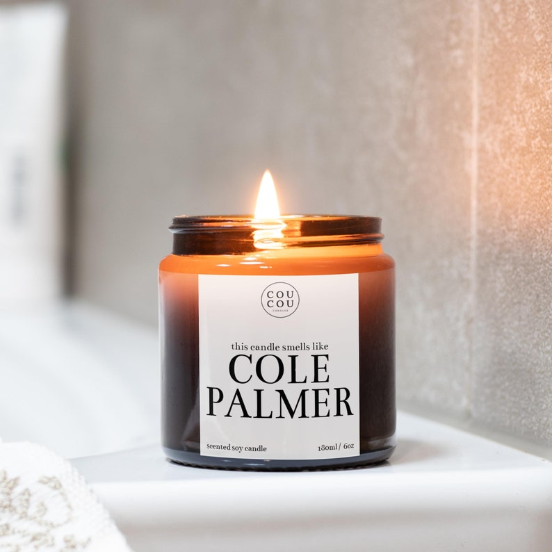 May include: A brown glass candle jar with a black label that reads "this candle smells like COLE PALMER" and "scented soy candle 180ml / 6oz". The candle is lit and the flame is visible.