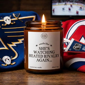 May include: A lit amber glass jar candle with a white label that reads "COU COU", "smells like", "SHE'S WATCHING HEATED RIVALRY AGAIN...", and "180ml/6oz". The candle is positioned in front of a blue hockey jersey and a red hockey jersey.