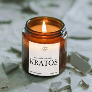 May include: A scented soy candle in a brown glass jar with a white label that reads Cou Cou, this candle smells like KRATOS, and 180ml/6oz. The candle features a lit wick with a warm yellow flame, casting a glow from within the amber glass container.