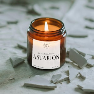 May include: A lit scented soy candle in a brown glass jar. The label reads "this candle smells like Astarion." The jar is 100ml / 6oz. The candle is surrounded by white, broken pieces. The flame is bright and the wax is a warm orange colour.