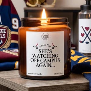 May include: A lit amber glass jar candle with a white label featuring the text "SMELLS LIKE SHE'S WATCHING OFF CAMPUS AGAIN..." and small hockey stick graphics. The label also displays the brand name "COU COU" and the volume 180ml / 6oz.