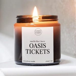 May include: A lit scented soy candle in a dark amber glass jar with a white label. The label features the text COU COU, smells like i have OASIS TICKETS, and 180ml / 6oz. The candle has a single burning wick with a bright flame at the centre.