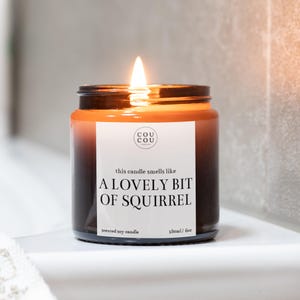 May include: A brown glass candle jar with a white label that reads "this candle smells like A LOVELY BIT OF SQUIRREL". The candle is lit and the flame is visible. The label also says "scented soy candle" and "180ml/6oz".