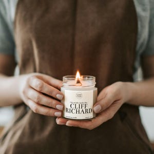 May include: A lit soy candle in a clear glass jar with a beige label. The label reads "This candle smells like Cliff Richard" and has the logo "Cou Cou" in a circle.