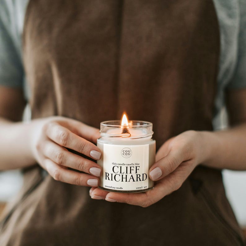 May include: A lit soy candle in a clear glass jar with a beige label. The label reads "This candle smells like Cliff Richard" and has the logo "Cou Cou" in a circle.
