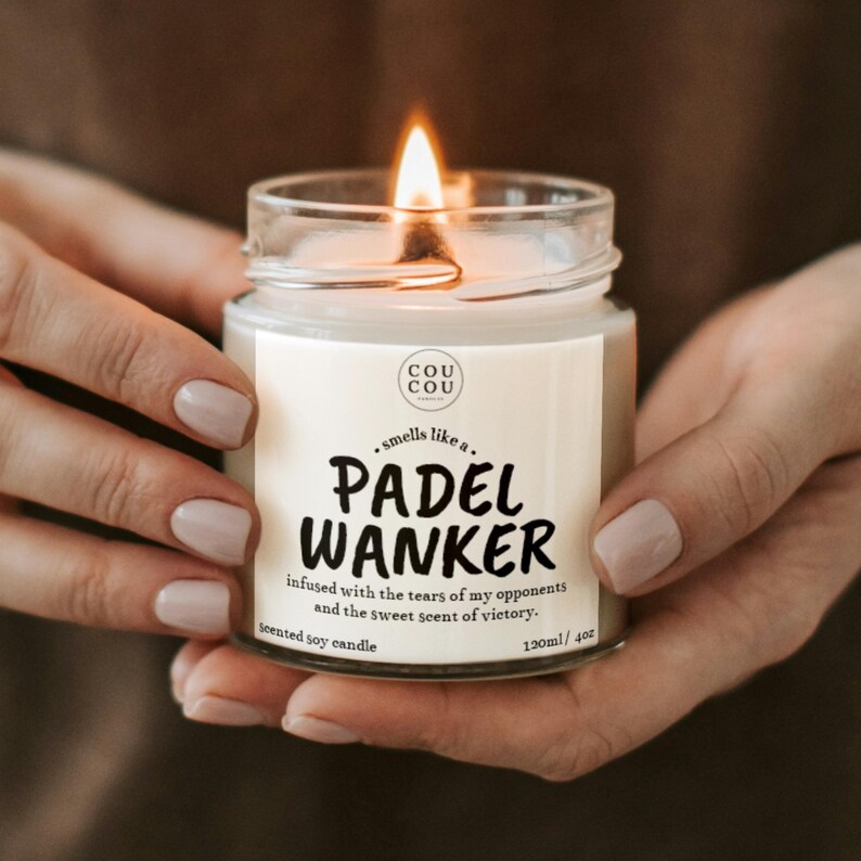 May include: A lit scented soy candle in a clear glass jar. The label reads "PADEL WANKER" with the text "infused with the tears of my opponents and the sweet scent of victory." The candle is 4 oz.