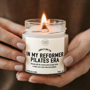 May include: A clear glass jar candle with a lit wick and a white label. The label features the text 'COU COU', 'SMELLS LIKE I'M IN MY REFORMER PILATES ERA', and 'infused with the sweet aroma of grip socks, a shaky core, and a few more pulses' with 120ml/4oz.