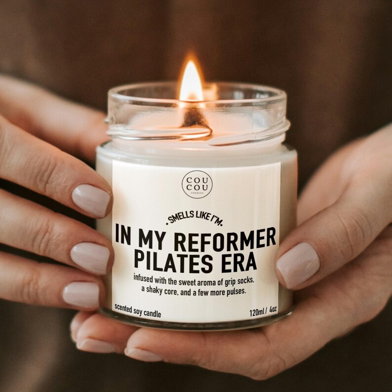 May include: A clear glass jar candle with a lit wick and a white label. The label features the text 'COU COU', 'SMELLS LIKE I'M IN MY REFORMER PILATES ERA', and 'infused with the sweet aroma of grip socks, a shaky core, and a few more pulses' with 120ml/4oz.
