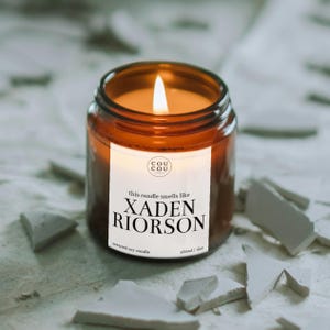 May include: A lit soy candle in an amber glass jar with a white label that reads "COU COU", "this candle smells like XADEN RIORSON", and "150ml / 6oz". The candle features a single burning wick and is filled with a warm, golden-toned wax.