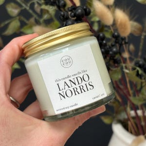May include: A glass candle jar with a gold lid. The label reads "this candle smells like LANDO NORRIS" and "scented soy candle 120ml / 4oz".
