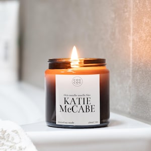 May include: A brown glass candle jar with a white label that reads "this candle smells like KATIE MCCABE scented soy candle 180ml / 6oz" and a smaller white label with the text "cou cou candles".