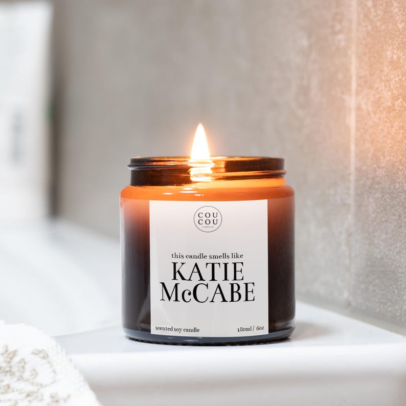 May include: A brown glass candle jar with a white label that reads "this candle smells like KATIE MCCABE scented soy candle 180ml / 6oz" and a smaller white label with the text "cou cou candles".