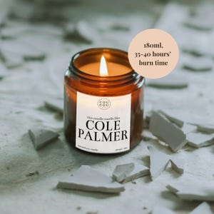 May include: A brown glass candle with a white label that reads "this candle smells like COLE PALMER". The candle is lit and the flame is visible. The candle is sitting on a white surface with broken pieces of white tile.