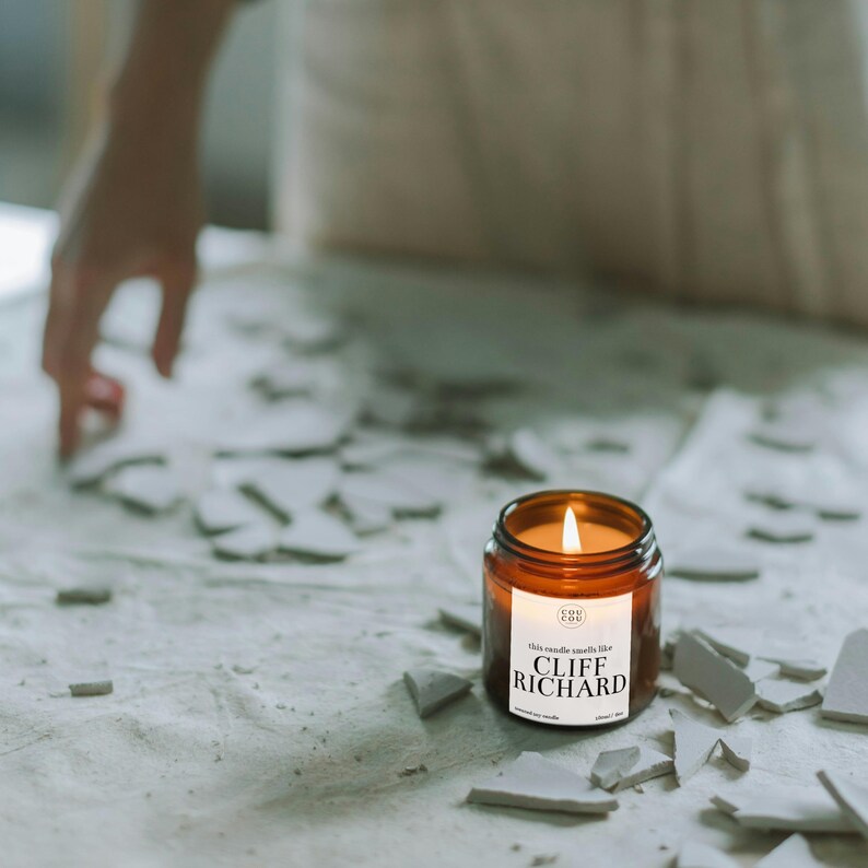 May include: A brown glass candle with a white label that reads "This candle smells like Cliff Richard". The candle is lit and sitting on a white surface with broken pieces of white tile.