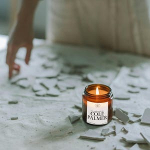 May include: A brown glass candle with a white label that reads "This candle smells like COLE PALMER". The candle is lit and sitting on a white surface with broken pieces of white tile.