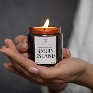 May include: A brown glass candle jar with a white label that reads "this candle smells like BARRY ISLAND" and "Cou Cou" in a smaller font. The candle is lit and the flame is visible.
