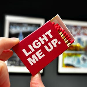 May include: A rectangular red matchbox with the white text LIGHT ME UP. printed on the front. The box is partially open, revealing a row of wooden matches with red tips arranged inside. The design is simple, featuring bold lettering.
