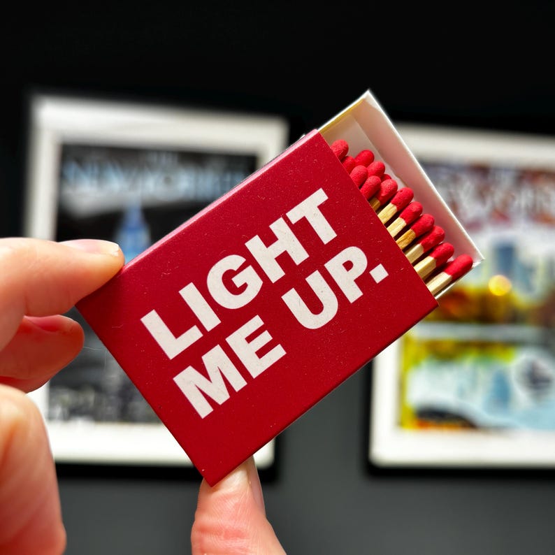May include: A rectangular red matchbox with the white text LIGHT ME UP. printed on the front. The box is partially open, revealing a row of wooden matches with red tips arranged inside. The design is simple, featuring bold lettering.