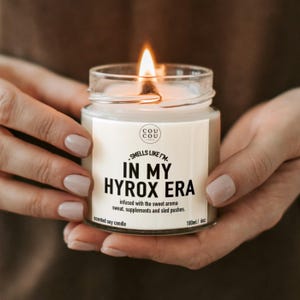 May include: A clear glass jar containing a scented soy candle with a lit wick. The white label features the text 'COU COU', 'SMELLS LIKE I'M IN MY HYROX ERA', and 'infused with the sweet aroma sweat, supplements and sled pushes.' It is 180ml/6oz.