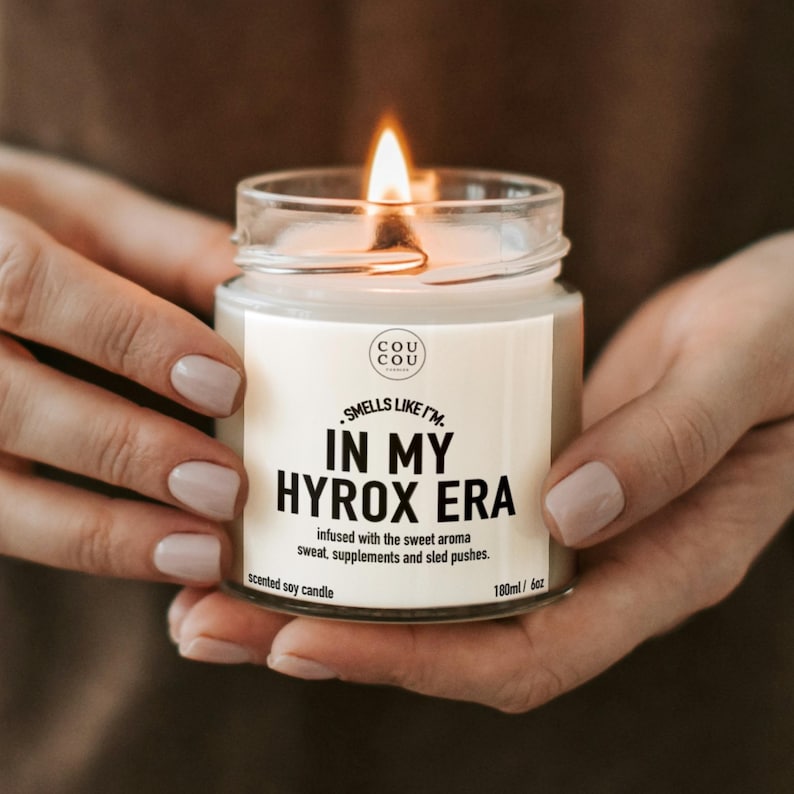 May include: A clear glass jar containing a scented soy candle with a lit wick. The white label features the text 'COU COU', 'SMELLS LIKE I'M IN MY HYROX ERA', and 'infused with the sweet aroma sweat, supplements and sled pushes.' It is 180ml/6oz.
