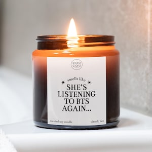 May include: A lit scented soy candle in a dark amber glass jar with a white label. The label features the text "COU COU CANDLES", "smells like", "SHE'S LISTENING TO BTS AGAIN...", "SCENTED SOY CANDLE", and "180ml / 6oz" in black serif and sans-serif fonts.