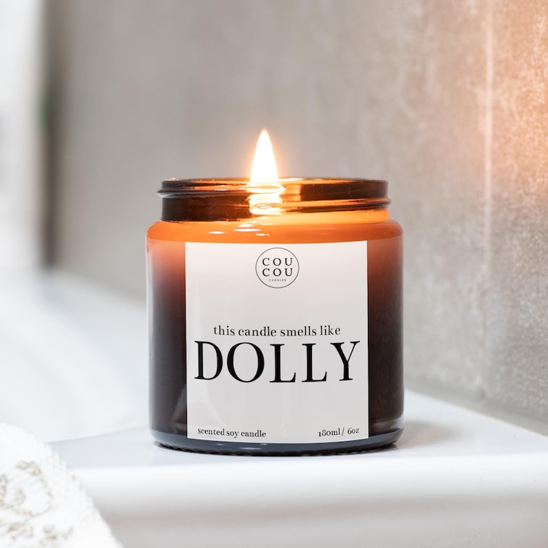 May include: A brown glass candle jar with a white label that reads "this candle smells like DOLLY" and "scented soy candle 180ml / 6oz". The candle is lit and the flame is visible.
