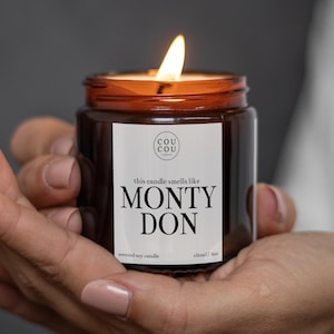 May include: A lit scented soy candle in an amber glass jar with a white label. The label features the text Cou Cou Candles, this candle smells like MONTY DON, and 180ml/6oz. The candle has a visible flame and a dark brown glass container.