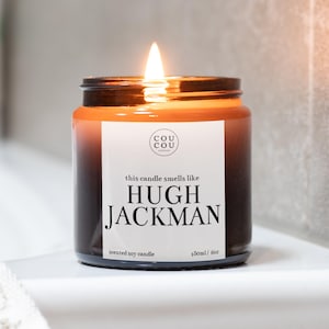 May include: A brown glass candle jar with a white label that reads "this candle smells like HUGH JACKMAN" and "scented soy candle 180ml / 6oz". The candle is lit and the flame is visible.