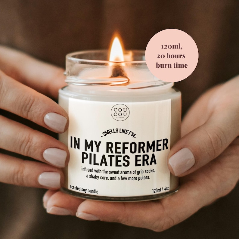 May include: A clear glass jar candle with a white label featuring the text "IN MY REFORMER PILATES ERA" and "120ml, 20 hours burn time". The candle has a lit wick and contains soy wax. The label also includes the brand name "COU COU" and descriptive text.