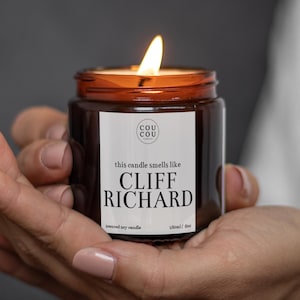 May include: A brown glass candle jar with a white label that reads "this candle smells like CLIFF RICHARD" and "scented soy candle 180ml / 6oz". The candle is lit and the flame is visible.