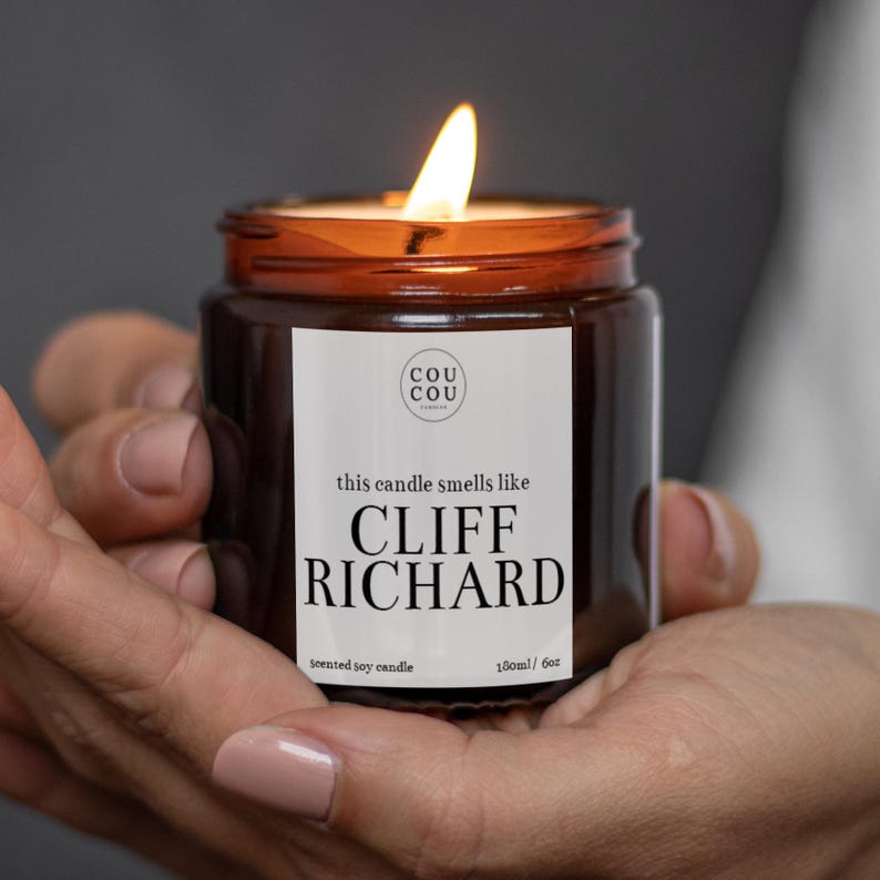 May include: A brown glass candle jar with a white label that reads "this candle smells like CLIFF RICHARD" and "scented soy candle 180ml / 6oz". The candle is lit and the flame is visible.