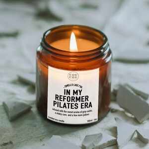 May include: A lit scented soy candle in an amber glass jar with a white label. The label reads: COU COU, SMELLS LIKE I'M IN MY REFORMER PILATES ERA, infused with the sweet aroma of grip socks, a shaky core, and a few more pulses, 180ml / 6oz.