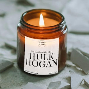 May include: A lit scented soy candle in a brown glass jar. The label reads "this candle smells like HULK HOGAN" with the brand logo and "180ml / 6oz". The candle is on a light-coloured surface.