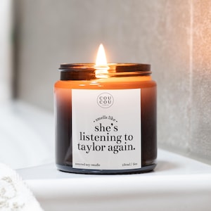 May include: A lit scented soy candle in a dark amber glass jar with a white label. The label features the text 'COU COU', 'smells like', 'she's listening to taylor again.', 'SCENTED SOY CANDLE', and '180ml / 6oz' in black, sans-serif typography.