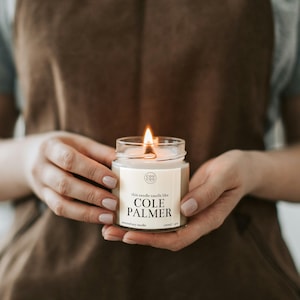 May include: A lit soy candle in a clear glass jar with a beige label that reads "this candle smells like COLE PALMER" and "cou cou" in a circle. The candle is being held by a person wearing a brown suede apron.