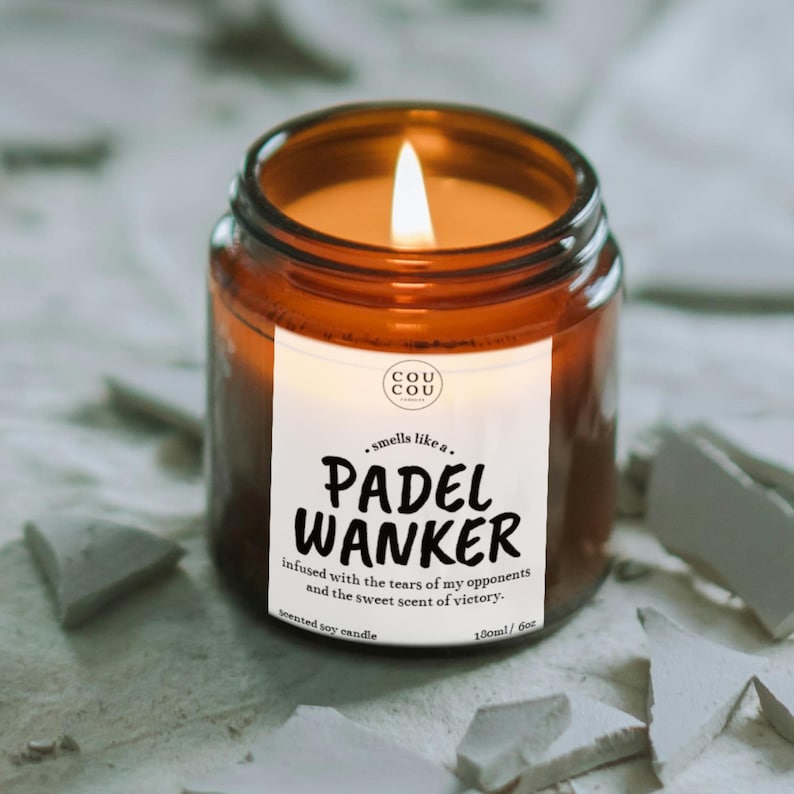 May include: A lit scented soy candle in a brown glass jar. The label reads "PADEL WANKER" with the text "smells like a..." above and "infused with the tears of my opponents and the sweet scent of victory" below. The candle is 100ml / 6oz.