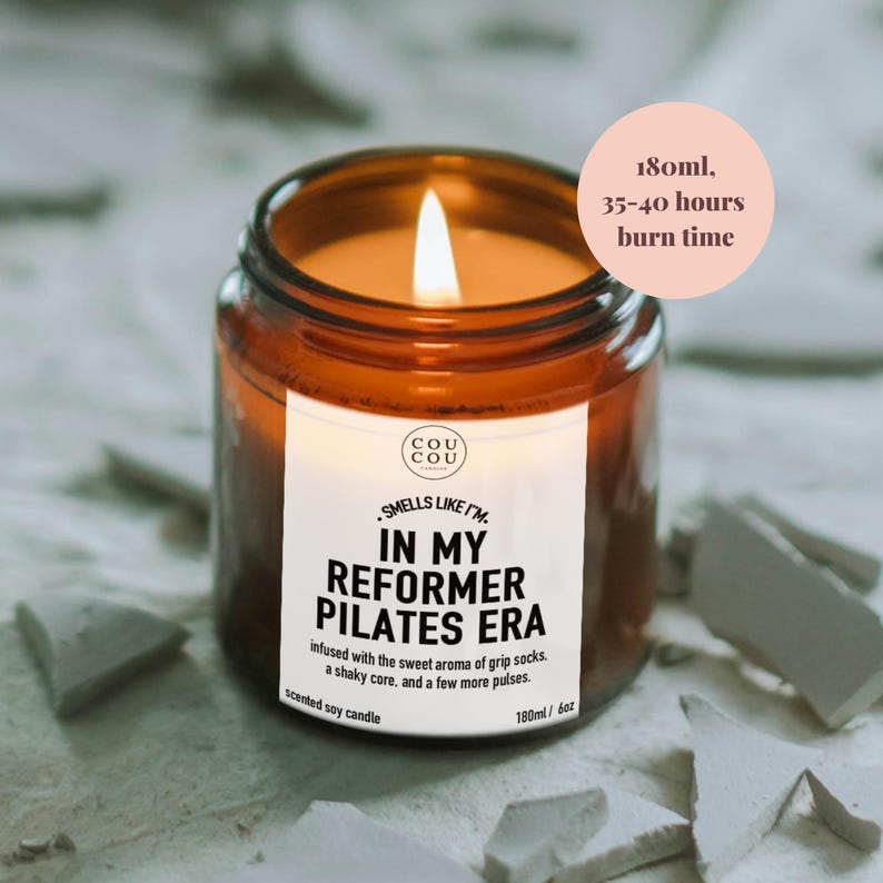 May include: A lit scented soy candle in an amber glass jar with a white label reading Cou Cou, Smells Like I'm In My Reformer Pilates Era, infused with the sweet aroma of grip socks, a shaky core, and a few more pulses. It is 6oz, 180ml, 35-40 hours burn time.