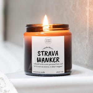 May include: A scented soy candle in a dark amber glass jar with a lit wick. The white label features the text 'COU COU', 'smells like a STRAVA WANKER', 'infused with a new personal best, and if it's not on strava, it didn't happen', '180ml/ 6oz'.