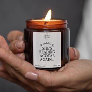 May include: A lit soy candle in a brown glass jar with a white label that reads Cou Cou, smells like SHE'S READING ACOTAR AGAIN, and 180ml/6oz. The candle features a bright flame and is contained within a cylindrical jar with a black screw-top lid.