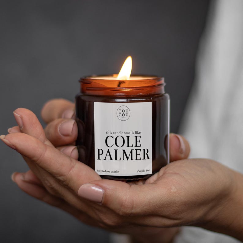 May include: A brown glass candle jar with a white label that reads "this candle smells like COLE PALMER" and "scented soy candle 120ml / 6oz". The candle is lit and the flame is visible.