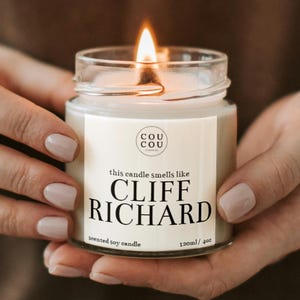 May include: A white soy candle in a clear glass jar with a black label that reads "this candle smells like CLIFF RICHARD", "scented soy candle", and "120ml / 4oz". The candle is lit and the flame is visible.