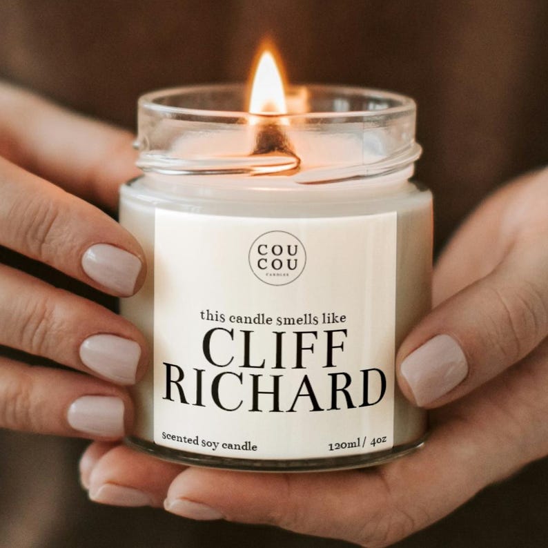 May include: A white soy candle in a clear glass jar with a black label that reads "this candle smells like CLIFF RICHARD", "scented soy candle", and "120ml / 4oz". The candle is lit and the flame is visible.