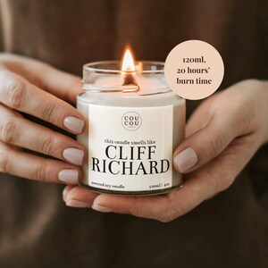 May include: A white soy candle in a glass jar with a black label that reads "this candle smells like CLIFF RICHARD" and "120ml/4oz scented soy candle". The candle is lit and the jar is being held by hands. A peach-colored circle with black text reads "120ml, 20 hours' burn time".