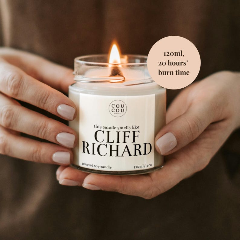 May include: A white soy candle in a glass jar with a black label that reads "this candle smells like CLIFF RICHARD" and "120ml/4oz scented soy candle". The candle is lit and the jar is being held by hands. A peach-colored circle with black text reads "120ml, 20 hours' burn time".