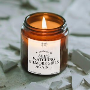 May include: A lit scented soy candle in an amber glass jar with a white label that reads Cou Cou, smells like SHE'S WATCHING GILMORE GIRLS AGAIN..., and 180ml / 6oz. The candle features a single burning wick with a warm orange glow inside the jar.