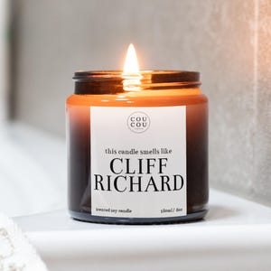May include: A brown glass candle jar with a white label that reads "this candle smells like CLIFF RICHARD" and "scented soy candle 180ml/6oz". The candle is lit and the flame is visible.