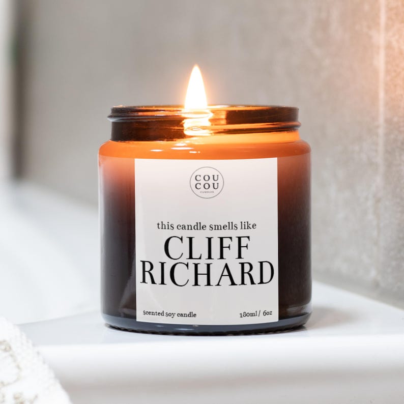 May include: A brown glass candle jar with a white label that reads "this candle smells like CLIFF RICHARD" and "scented soy candle 180ml/6oz". The candle is lit and the flame is visible.