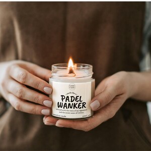 May include: A lit scented soy candle in a clear glass jar. The label reads "PADEL WANKER" with additional text. The candle is held in two hands. The candle is 4 oz.