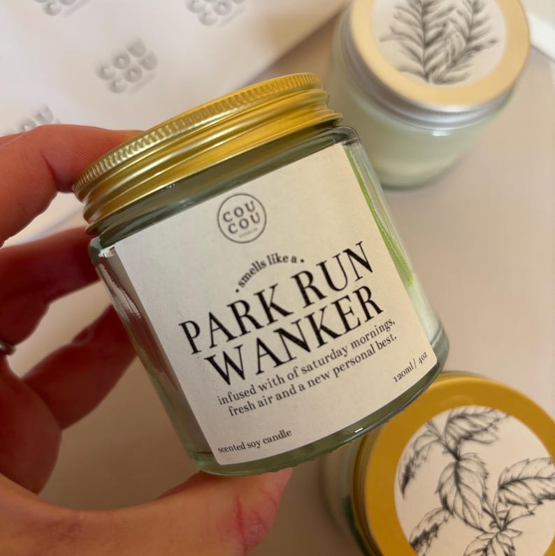 May include: A clear glass jar with a gold lid, labeled "PARK RUN WANKER" with the text "smells like a... infused with of saturday mornings, fresh air and a new personal best." The jar contains a scented soy candle, 4 oz.