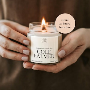 May include: A white soy candle in a glass jar with a black label that reads "this candle smells like COLE PALMER" and "120ml/4oz". The candle is lit and has a flame. A white circle with black text reads "120ml, 20 hours' burn time".